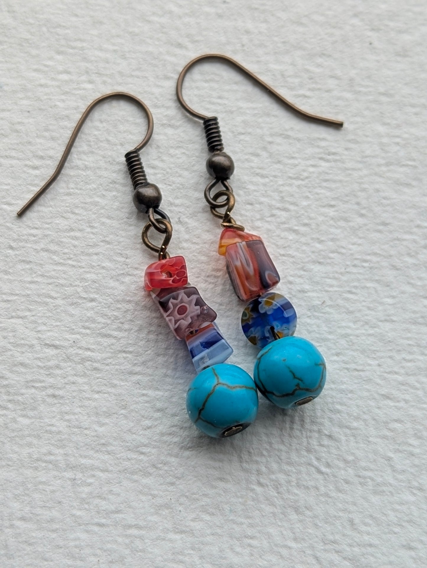 Handmade dangle earrings featuring colorful millefiori glass beads and round, light blue beads with a veined pattern, resembling turquoise. Finished with bronze-tone ear hooks.