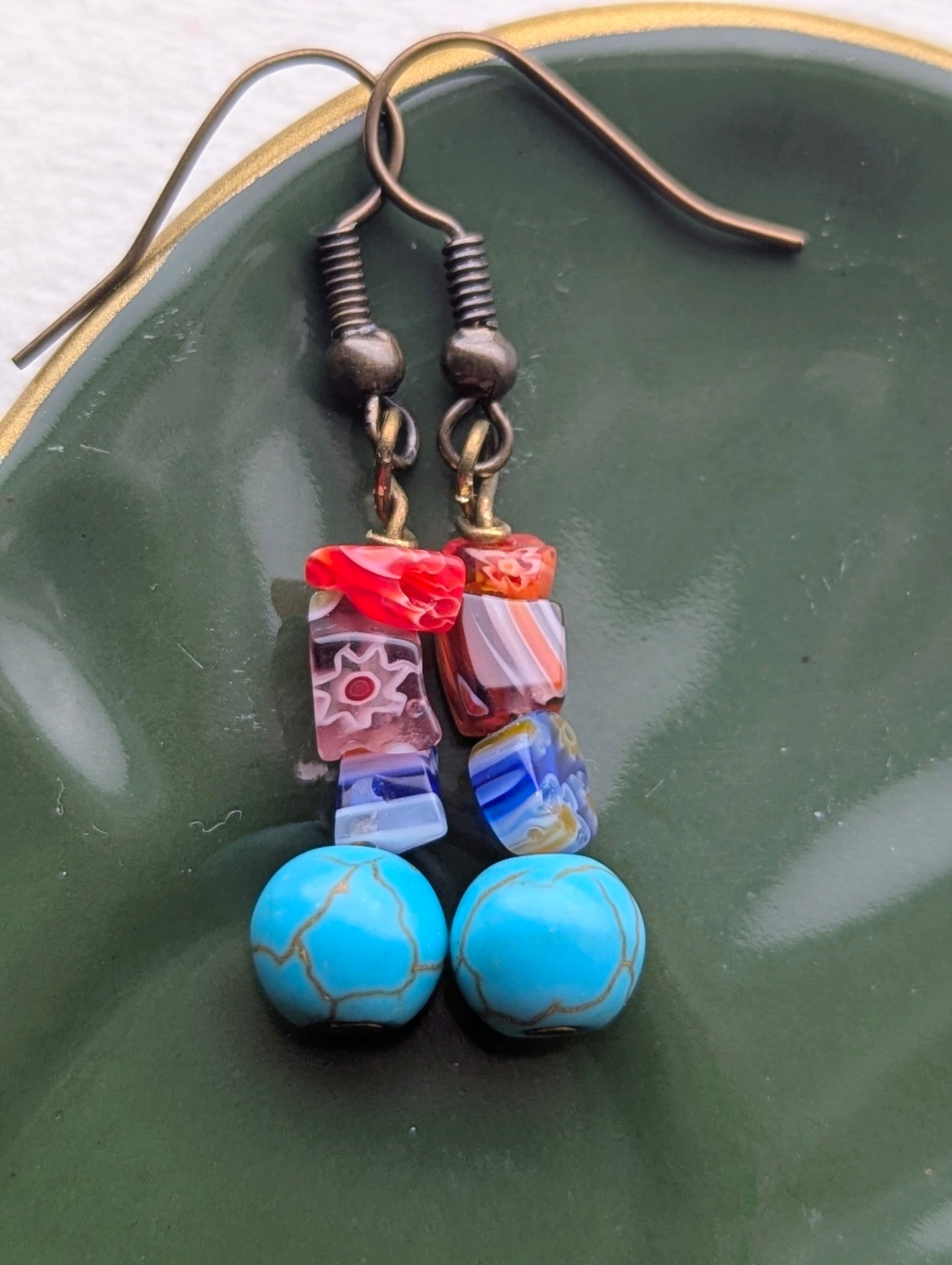 Handmade dangle earrings featuring colorful millefiori glass beads and round, light blue beads with a veined pattern, resembling turquoise. Finished with bronze-tone ear hooks.